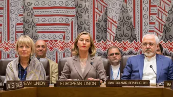 L'image montre une réunion internationale avec plusieurs personnes assises à une table. On peut voir des panneaux identifiant les différents participants, tels que l'Union européenne et l'Iran. Trois figures centrales semblent être des représentants importants, avec des expressions sérieuses. En arrière-plan, il y a des individus qui écoutent attentivement. Le décor est riche et coloré, avec des motifs géométriques sur le mur derrière eux, suggérant un cadre formel pour des discussions diplomatiques.