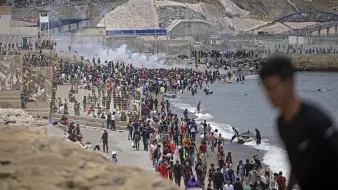 L'image montre une foule nombreuse sur une plage, avec des personnes rassemblées près de l'eau et d'autres sur le rivage. En arrière-plan, on peut voir des collines ainsi que des structures de béton. Il y a également des éléments de chaos, comme de la fumée, ce qui pourrait indiquer une situation tendue ou un événement significatif. Des gens semblent se déplacer, certains en direction de la mer, tandis que d'autres se trouvent sur le sable ou dans l'eau.