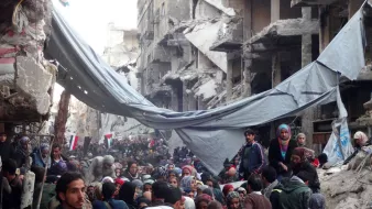 L'image montre une scène de destruction massive dans une zone urbaine, probablement après un conflit ou une catastrophe. On peut voir des bâtiments gravement endommagés, avec des murs effondrés et des débris partout. Une foule de personnes, dont certaines semblent déplacées, se rassemble dans la rue. Il y a des tentes et des bâches qui servent d'abris temporaires. Des membres d'organisations humanitaires, identifiables par leurs vêtements distinctifs, sont présents pour aider. L'ambiance générale est celle de la détresse et de la recherche d'aide dans une situation de crise.