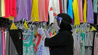 L'image montre une personne dans un magasin de vêtements, portant un hijab et un écran facial. Elle porte des gants et examine des vêtements accrochés sur des cintres. Les étagères à proximité sont remplies de vêtements colorés, principalement des t-shirts et des leggings, dans des teintes vives comme le violet, le jaune et le vert. L'ambiance semble être celle d'un environnement de shopping où l'on cherche des articles vestimentaires.
