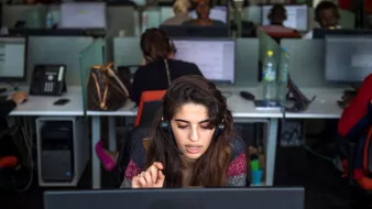 L'image montre un bureau avec plusieurs employés travaillant sur des ordinateurs. Au premier plan, une jeune femme porte un casque et semble concentrée sur son écran. Elle gesticule avec une main, probablement en train de communiquer avec un client. En arrière-plan, d'autres personnes sont visibles, toutes occupées à leurs tâches. L'environnement a l'air moderne, avec des bureaux en open space et des équipements de travail.