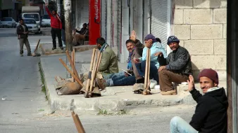 L'image montre un groupe d'hommes assis sur le bord de la route, apparemment en attente. Ils semblent discuter et certains saluent la caméra. L'environnement a l'air urbain, avec des bâtiments en arrière-plan et une rue. Des outils de travail sont visibles, suggérant qu'ils pourraient être des travailleurs. L'atmosphère semble décontractée malgré le cadre urbain.