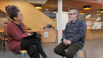 L'image montre deux personnes assises dans un espace intérieur. À gauche, une femme avec des cheveux bouclés et des lunettes, vêtue d'un pull rouge, tient une tablette. À droite, un homme portant des lunettes et une chemise bleu foncé est assis, les mains sur ses genoux. L'environnement est moderne, avec des murs en bois et des éléments de décoration qui donnent une ambiance chaleureuse. Cela semble être une interview ou une discussion.