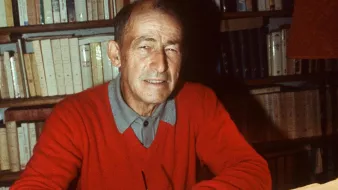 L'image montre un homme assis à un bureau, souriant, en train d'écrire sur une feuille de papier avec un stylo. Il porte un pull rouge et une chemise à col. En arrière-plan, on peut voir une bibliothèque remplie de livres. Sur le bureau, il y a également des feuilles de papier, peut-être des notes ou un manuscrit en cours de rédaction. L'éclairage crée une ambiance chaleureuse et studieuse.