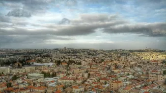 L'image montre une vue panoramique d'une ville, probablement Jérusalem, avec des bâtiments disposés sur plusieurs collines. Les toits sont principalement en tuiles rouges, et il y a de nombreux édifices en pierre. Le ciel est nuageux, ajoutant à l'ambiance de la scène, avec une lumière douce qui éclaire la ville. On peut également apercevoir des zones de verdure entre les bâtiments, ce qui ajoute de la profondeur au paysage urbain.