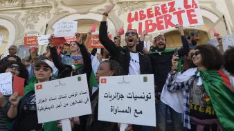 L'image montre un groupe de personnes rassemblées lors d'une manifestation. Les manifestants portent des pancartes et des drapeaux, exprimant des slogans tels que "Libérez la liberté". Les participants semblent engagés et passionnés, brandissant des affiches avec des messages en arabe et en français, appelant à la justice et à l'égalité. L'ambiance est énergique et déterminée, témoignant d'une dynamique de revendication sociale ou politique. L'arrière-plan présente une architecture caractéristique, ajoutant au contexte de l'événement.
