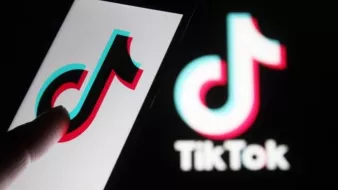 L'image montre un smartphone avec le logo de TikTok affiché à l'écran. On peut voir un doigt en train de toucher ou interagir avec le logo. À l'arrière-plan, il y a le logo de TikTok dans un style similaire, mais peut-être moins détaillé. Les couleurs principales du logo sont le noir, le rouge et le bleu, créant un effet visuel attrayant.