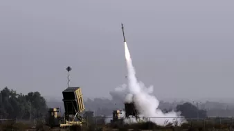 L'image montre un système de défense antimissile en action, avec un missile tiré vers le ciel. On peut voir les lanceurs de missiles, qui sont situés sur un terrain ouvert, entourés de végétation. Une traînée de fumée s'élève du lanceur, indiquant le moment du lancement. Le ciel est clair, avec une légère brume qui lui confère une atmosphère chargée.