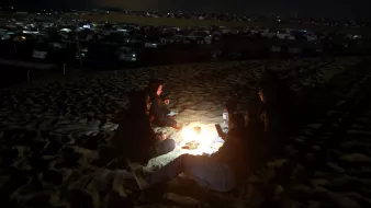 L'image montre un groupe de personnes assises autour d'un feu de camp sur une dune de sable, en pleine nuit. Ils semblent se réchauffer et passer un moment ensemble. En arrière-plan, on peut apercevoir des tentes et d'autres structures, suggérant qu'ils se trouvent dans un campement. La lumière du feu éclaire leurs visages et crée une atmosphère chaleureuse malgré l'obscurité environnante.