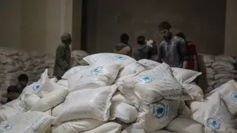 L'image montre une pièce sombre remplie de sacs de nourriture, probablement de farine ou de riz, empilés sur le sol. À l'arrière-plan, plusieurs personnes sont présentes, mais leurs visages ne sont pas clairement visibles. Les sacs portent des inscriptions, indiquant probablement qu'ils contiennent de l'aide alimentaire. L'atmosphère semble être celle d'une distribution ou d'un entreposage de secours.