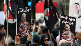 L'image montre une manifestation avec des personnes tenant des pancartes et des drapeaux. Les pancartes contiennent des portraits de personnes et des textes en arabe, évoquant des messages politiques ou sociaux. La foule semble engagée dans une cause, exprimant des émotions fortes. On peut voir une diversité de participants, certains portant des vêtements traditionnels et d'autres plus modernes, ainsi qu'un fond urbain suggérant un rassemblement public.