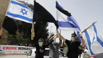 L'image montre un groupe de personnes en train de manifester. Elles tiennent des drapeaux, y compris le drapeau israélien et un autre drapeau aux couleurs noire et blanche. Tous les participants portent des masques sur le visage, ce qui suggère qu'ils prennent des précautions sanitaires. En arrière-plan, on peut voir une banderole qui semble contenir un message critique envers le gouvernement. L'atmosphère semble être celle d'une protestation pacifique.