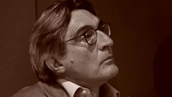 L'image montre un homme aux cheveux grisonnants, portant des lunettes et un costume clair. Il semble pensif, regardant quelque chose hors champ. L'éclairage et le ton de l'image lui donnent un aspect rétro, suggérant une ambiance contemplative.