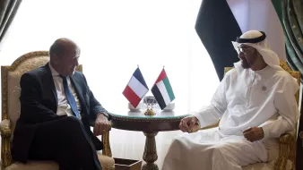 L'image montre deux hommes assis sur des fauteuils dans un cadre élégant. Ils discutent autour d'une petite table portant des drapeaux de la France et des Émirats Arabes Unis. L'un des hommes est vêtu d'un costume, tandis que l'autre porte une tenue traditionnelle blanche. Leurs expressions semblent amicales et engageantes, suggérant une conversation diplomatique. L'arrière-plan est décoré de rideaux et d'autres éléments luxueux.