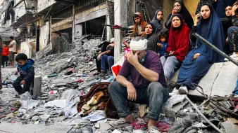 L'image montre un groupe de personnes assises sur des décombres dans une zone de conflit. Au premier plan, un homme, la tête bandée, exprime une grande tristesse ou du désespoir, tandis qu'à proximité, plusieurs femmes en vêtements sombres regardent vers l'objectif, certaines avec une expression préoccupée. Un petit garçon est assis un peu plus loin, semblant perdu dans ses pensées. L'environnement est chaotique, avec des ruines et des débris autour, suggérant des dommages importants causés par des événements récents. L'atmosphère générale est lourde, reflétant la douleur et la résilience des personnes présentes.