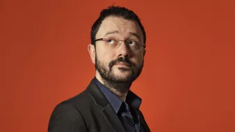 L'image montre un homme vêtu d'un costume noir sur une chemise bleu foncé, regardant légèrement sur le côté. Il a des lunettes, une barbe et un léger sourire. L'arrière-plan est orange, ce qui crée un contraste vif et dynamique avec sa tenue sombre. L'expression de son visage semble pensif ou intrigué.