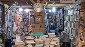 L'image montre une librairie pleine de livres, avec des étagères qui montent jusqu'au plafond. Il y a une grande horloge murale qui indique l'heure. Au centre, des piles de livres sont empilées sur une table, tandis qu'un homme est penché sur une étagère, en train de chercher ou de réorganiser des livres. L'ambiance semble chaleureuse et accueillante, avec un éclairage doux provenant de lampes suspendues. Une inscription en arabe est visible au mur.