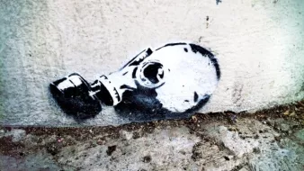 L'image montre un graffiti représentant une tête de mort portant un masque à gaz. Ce dessin est peint en noir et blanc et est appliqué sur un mur, près du sol. Le contraste entre le noir et le blanc met en évidence les contours du masque et de la tête, créant une image saisissante et inquiétante. Le style est à la fois moderne et urbain, typique des œuvres de street art.