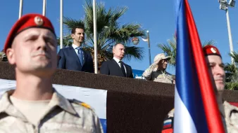 L'image montre un groupe de personnes, dont deux figures de haut rang, sur une plateforme en extérieur, avec un fond de palmiers et un ciel bleu. Au premier plan, des soldats en uniforme, probablement d'une unité militaire, sont présents, portant des drapeaux. Les expressions des personnes sur la plateforme semblent sérieuses et concentrées, indiquant un événement officiel ou militaire.