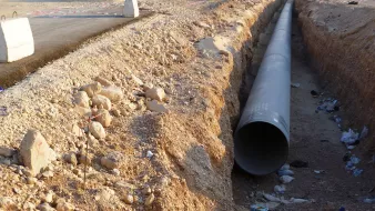 L'image montre un espace de construction où un grand tuyau en métal est disposé dans une tranchée creusée dans le sol. Le terrain environnant est aride et rocailleux, avec quelques débris, comme des bouteilles, dispersés dans la tranchée. Au fond, on peut voir des engins de chantier et une route partiellement construite. L'ambiance générale évoque un projet d'infrastructure en cours.