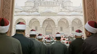 L'image montre un grand groupe de personnes, probablement des religieux ou des fidèles, qui se tiennent debout, tournés vers un imposant bâtiment architectural qui semble être une mosquée. Les hommes portent des vêtements similaires, souvent dans des tons sombres, et certains portent des turbans ou des chapeaux à pompons rouges. L'ambiance semble solennelle et respectueuse, suggérant un moment de prière ou de rassemblement communautaire. Les détails architecturaux de la mosquée, avec ses coupoles et arches majestueuses, ajoutent à l'imposante scène.