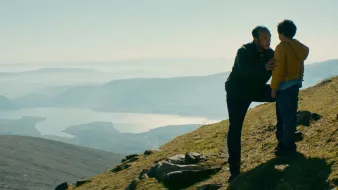 L'image montre un homme et un enfant sur une colline, face à un paysage naturel vaste et montagneux. L'homme semble parler à l'enfant, peut-être pour lui donner des conseils ou partager un moment d'intimité. En arrière-plan, on aperçoit un lac scintillant et des montagnes, le tout baigné par une lumière douce, ce qui crée une atmosphère sereine et contemplative. La scène évoque des thèmes de connexion et de découverte en pleine nature.