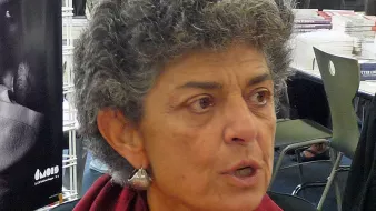 Je ne sais pas qui est la personne sur l'image. Cependant, je peux décrire l'apparence générale : il s'agit d'une femme avec des cheveux gris et bouclés, portant un gilet noir et une écharpe rouge. Elle semble engagée dans une conversation, exprimant des émotions. En arrière-plan, on distingue des personnes et des livres.