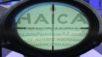 L'image montre un réticule de visée, typique d'une lunette de tir, qui se superpose à un fond avec le mot "HAICA" visible. Ce mot est associé à une autorité indépendante en matière de communication auditive, peut-être en rapport avec la régulation des médias. Le contraste entre l'optique et le texte suggère un thème de surveillance ou de contrôle. Le fond semble comporter divers éléments, peut-être liés à des événements ou des institutions médiatiques.