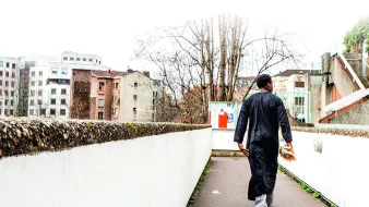 L'image montre un homme marchant sur un chemin urbain. Il porte une tenue traditionnelle, une sorte de longue robe noire. En arrière-plan, on peut voir des bâtiments modernes et anciens, ainsi que des arbres dépouillés. L'environnement semble calme, avec une lumière diffuse, peut-être un jour nuageux. L'homme avance vers l'horizon, de dos, ce qui donne une impression de solitude et de contemplation.