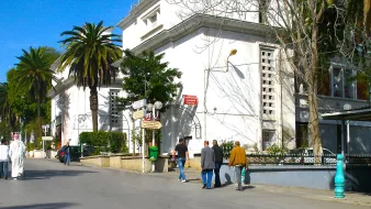 L'image montre une rue dégagée avec des bâtiments blancs et modernes de chaque côté. Des palmiers bordent le chemin, ajoutant une touche tropicale à l'environnement. On peut voir plusieurs personnes marchant le long de la rue, certaines en tenue traditionnelle. Le ciel est bleu et ensoleillé, ce qui donne une ambiance agréable et décontractée à la scène. Les façades des bâtiments présentent une architecture typique de la région, avec des détails subtils comme des lampadaires et des panneaux indiquant des directions.