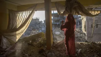 L'image montre une scène à l'intérieur d'un bâtiment endommagé. On aperçoit une femme vêtue d'un vêtement traditionnel, se tenant près d'une fenêtre qui donne sur des ruines. De l'autre côté, on voit des décombres de bâtiments effondrés, laissant entrevoir la désolation d'un environnement détruit. La lumière du soleil filtre à travers les rideaux, ajoutant une ambiance à la fois triste et poignante à la scène.