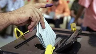 L'image montre une main avec un doigt marqué de violet, symbole d'un vote. La personne insère un bulletin de vote dans une urne électorale. En arrière-plan, on peut voir d'autres individus, probablement en train d'attendre ou de voter eux aussi. L'atmosphère semble sérieuse et engageante, illustrant un moment clé du processus démocratique.