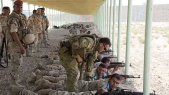 L'image montre un groupe de soldats en train de s'entraîner sur un champ de tir, allongés à terre avec leurs armes. Certains soldats portent des uniformes beiges avec des motifs camouflés, tandis qu'un instructeur semble donner des conseils à un autre soldat. En arrière-plan, on aperçoit d'autres soldats attendant ou surveillant l'exercice. L'environnement est désertique avec une structure en métal au-dessus d'eux, offrant un abri contre le soleil.