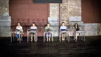 L'image montre un groupe de cinq personnes assises sur des chaises, disposées en ligne. Elles portent des vêtements simples et sont pieds nus, ce qui donne une impression de simplicité et d'intimité. L'environnement est industriel, avec des murs en brique apparente et une lumière tamisée qui crée une ambiance à la fois contemplative et moderne. Les personnes semblent concentrées, chacune avec un objet devant elle, suggérant un moment de performance ou de réflexion partagée.