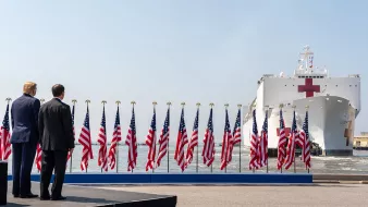 L'image montre une scène avec deux hommes de dos, face à un grand navire hospitalier qui arrive. Le navire est identifiable grâce à sa croix rouge sur le côté. Au premier plan, un podium est visible, entouré de multiples drapeaux américains flottant au vent. Le ciel est clair et lumineux, suggérant une journée ensoleillée. L'atmosphère semble solennelle, probablement en lien avec un événement officiel ou une cérémonie.