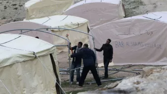 L'image montre plusieurs tentes de type campement, qui semblent être installées dans un environnement extérieur. Les tentes ont des écrits en arabe et en anglais, mentionnant "BAB AL SHAMS VILLAGE". On observe également des individus en train de travailler ensemble pour monter les structures des tentes. L'ensemble de la scène évoque des activités de construction ou d'aménagement dans un cadre probablement temporaire.