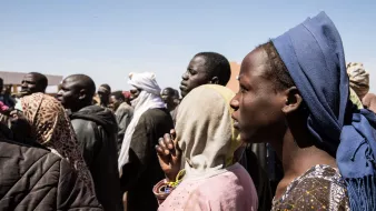L'image montre un groupe de personnes, probablement en attente ou rassemblées pour une raison quelconque. On peut voir des visages divers avec des expressions sérieuses. Certaines personnes portent des vêtements traditionnels et des accessoires variés, comme des écharpes ou des foulards. L'arrière-plan est flou, mais le ciel est dégagé. L'ambiance semble concentrée, peut-être en lien avec un événement important.