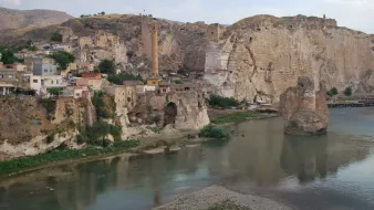 L'image montre un paysage pittoresque avec des formations rocheuses impressionnantes en arrière-plan. On y voit un village construit sur les flancs de la montagne, avec des maisons aux couleurs chaudes. Un fleuve calme coule au premier plan, reflétant les alentours. Quelques arbres verdoyants et des ruines anciennes ajoutent à la beauté historique de la scène. L'atmosphère est tranquille, dominée par les teintes naturelles des roches et de l'eau.