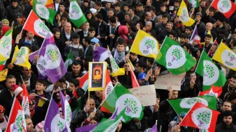 L'image montre une foule de manifestants brandissant des drapeaux colorés, principalement des couleurs violet, vert et rouge, qui semblent appartenir à un mouvement politique. Certaines personnes portent des affiches ou des pancartes. L'atmosphère semble dynamique et engagée, avec une concentration sur la participation collective. Les visages des manifestants sont variés et montrent un mélange de détermination et d'unité. Les drapeaux, avec des symboles, suggèrent une forte identité politique.