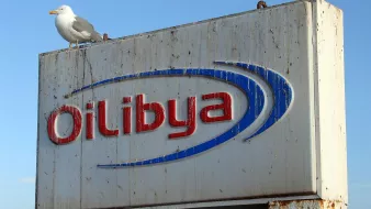 L'image montre un panneau publicitaire corrodé et usé portant le nom "Oil Libya" en lettres bleues et rouges. Au sommet du panneau, un goéland est posé. En arrière-plan, le ciel est clair, suggérant une belle journée. Le panneau semble être situé près de l'eau, probablement dans un port ou une zone côtière.