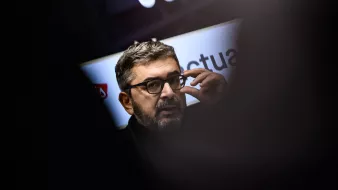 L'image montre un homme portant des lunettes et ayant une barbe courte. Il semble engagé dans une conversation ou une présentation, en touchant son visage avec une main. En arrière-plan, des éléments graphiques ou des logos sont flous, ce qui suggère qu'il se trouve dans un cadre professionnel ou public. L'éclairage met en avant son visage, tandis que des silhouettes floues d'autres personnes sont visibles autour.