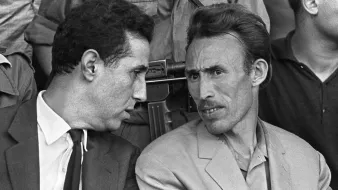 Cette image en noir et blanc montre deux hommes en discussion. L'un porte un costume sombre avec une cravate, tandis que l'autre est habillé de manière plus décontractée avec une veste claire. Ils semblent en train d'échanger des informations ou des réflexions, affichant des expressions attentives. En arrière-plan, on peut apercevoir d'autres personnes, et un fusil est visible, suggérant un contexte de tension ou de sécurité. L'image capte une moment d'intimité entre les deux hommes.