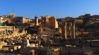 L'image montre un site archéologique avec des ruines anciennes, probablement d'un temple, entourées de colonnes et de blocs de pierre. En arrière-plan, on voit des bâtiments modernes et des structures urbaines s'élevant sous un ciel bleu. La lumière du soleil éclaire les ruines, créant un contraste entre l'ancien et le moderne. Ce lieu évoque une riche histoire et une importance culturelle.