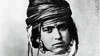 L'image montre un portrait noir et blanc d'une personne portant un turban orné et des bijoux. Le regard de la personne est intense, et son expression transmet une certaine dignité. Elle est vêtue de vêtements traditionnels qui reflètent une culture spécifique. Les détails du turban et des ornements ajoutent une dimension riche et texturée à l'image.