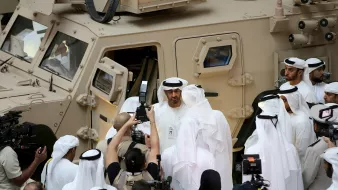 L'image montre un groupe de personnes, probablement des dignitaires, réunis autour d'un véhicule militaire dans un environnement formel. Plusieurs hommes portent des vêtements traditionnels, comme des kandouras et des ghutras. Il y a également des journalistes et des caméras en train de filmer ou de prendre des photos, ce qui suggère un événement médiatique ou une visite officielle. Le véhicule militaire est de couleur beige et semble robuste, avec plusieurs fenêtres et des équipements visibles.