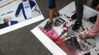 L'image montre des affiches au sol sur lesquelles sont piétinées des images de personnalités politiques, notamment Benjamin Netanyahu, Donald Trump et d'autres figures. Les affiches semblent être liées à une manifestation ou une protestation, avec des couleurs vives et des motifs qui attirent l'attention. Les personnes présentes dans l'image sont en train de marcher dessus, suggérant une attitude de rejet ou de mécontentement envers ces figures. Les tonnerres de couleurs et le contenu des affiches pourraient refléter des sentiments politiques forts.