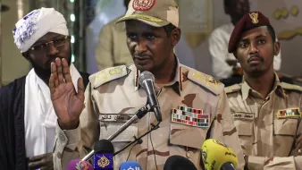 L'image montre un homme en uniforme militaire, portant une casquette décorée, qui s'exprime devant un micro lors d'une conférence de presse. Il semble être en train de faire une déclaration importante, avec une main levée. En arrière-plan, deux autres hommes sont également présents, l'un portant un chapeau traditionnel et l'autre en uniforme militaire. Divers microphones de médias sont placés devant lui. L'ambiance semble sérieuse, suggérant un contexte politique ou militaire.