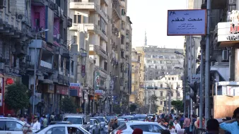 L'image montre une rue animée dans une ville, probablement une métropole. On peut voir de nombreux bâtiments de plusieurs étages qui bordent la route. Les voitures sont garées et circulent, créant un certain embouteillage. Une foule de personnes se déplace le long des trottoirs. Des panneaux de signalisation et des enseignes en arabe sont visibles, indiquant que cette scène se déroule dans un pays arabophone. L'atmosphère semble dynamique et vivante, typique d'une grande ville.