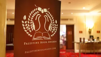 L'image représente un panneau de présentation pour les Palestine Book Awards. Ce panneau arbore un design avec des éléments graphiques, incluant une illustration d'un livre ouvert entouré de feuilles. Le texte indique "Palestine Book Awards" ainsi que le site web associé. L'arrière-plan semble être une salle de réception, avec un sol tapissé et des éclairages doux.