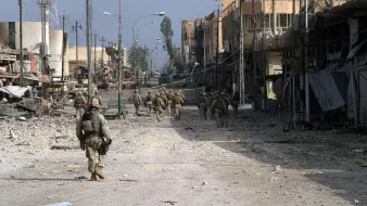 L'image montre une scène urbanisée dévastée, probablement dans une zone de conflit. On y voit des soldats marchant sur une rue déserte, entourés de bâtiments partiellement détruits. Le sol est jonché de débris, et l'atmosphère semble chargée de tension et de désolation. Les structures autour d'eux, avec des fils électriques en désordre, témoignent des ravages causés par le conflit. La lumière est diffuse, ce qui renforce le sentiment d'un environnement difficile et chaotique.